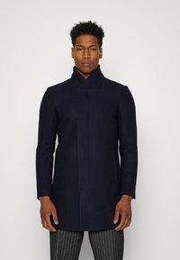 Abrigo de mezcla de lana color navy con un cuello alto, bolsillos frontal tipo parche y un ajuste entallado. La textura suave y el corte recto realzan su diseño moderno.