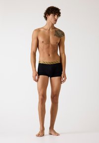 Young man with curly hair models black boxer briefs with gold patterned waistband, standing barefoot against a plain white background.