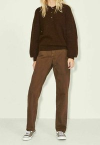 Brauner Strickpullover mit Rundhalsausschnitt und Dreiknopfleiste, kombiniert mit braunen Hosen mit weitem Bein und grauen Canvas-Schuhen.