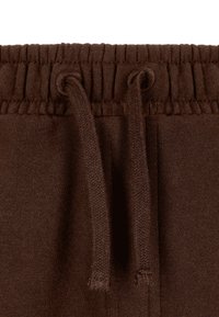 Bruine trekkoord tailleband van joggingbroek met geribbelde textuur en geweven accenten. Heeft twee trekkoorden met afgeronde uiteinden.