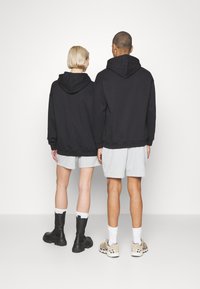 Two individuals wear matching black hoodies and gray shorts. The hoodies have a relaxed fit; one person wears black boots, the other sneakers.