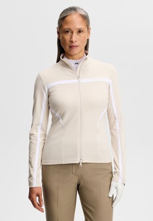 Femme portant une veste de sport beige à fermeture éclair avec des détails de bandes blanches, un pantalon couleur sable et un gant de golf blanc à la main gauche, sur un fond uni.