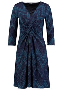 V-kaula-aukollinen mekko, jossa pitkät hihat ja sinivihreä siksak-kuvio. Ryppyinen yläosa ja liehuva helma, teksturoitu kankaan pinta, jossa kimallusta.