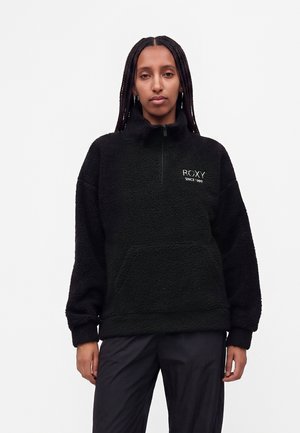 Donna con capelli intrecciati che indossa un pullover in pile nero con mezza cerniera e logo ROXY sul petto su sfondo bianco.