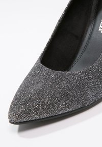 Chaussure plate noire pailletée avec un bout pointu et une doublure intérieure lisse. La tige texturée présente des paillettes métalliques, créant un effet scintillant.