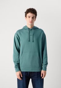 Felpa teal realizzata in materiale morbido, con tasca frontale, polsini a coste e cappuccio regolabile. Design semplice senza motivi o grafiche.