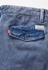 Blauwe ribstof met verticale strepen, een achterzak met een knoop en een rood Levi's logo etiket. Etiket zichtbaar met merkdetails.