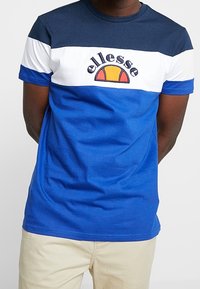 Muž v tričku s barevným blokem modré, bílé a námořnické barvy s logem "ellesse" v námořnické a oranžové, doplněný béžovými kalhotami.