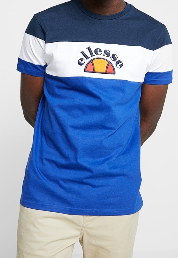 Muž v tričku s barevným blokem modré, bílé a námořnické barvy s logem "ellesse" v námořnické a oranžové, doplněný béžovými kalhotami.