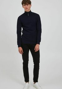 Pullover blu navy con texture a coste zigzag, colletto alto, chiusura a mezza zip; abbinato a pantaloni neri slim-fit e sneakers bianche.