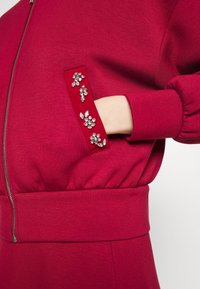 Czerwona bluza z kapturem na zamek ma teksturowany materiał i ściągacze, a kieszeń ozdobiona jest kwiatowymi zdobieniami z kamieni kryształowych.