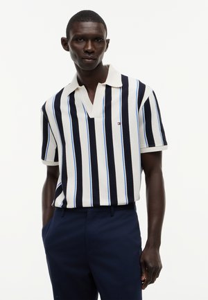 Mann trägt weißes Poloshirt mit schwarzen und blauen vertikalen Streifen und dunkelblaue Hose vor schlichtem weißem Hintergrund.