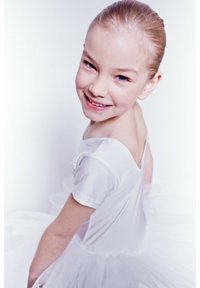 Weißes Ballettkleid mit transparenter Tüll-Rock, kurzen Ärmeln und einem tiefen Rückenausschnitt. Kind mit hellbraunem Haar, das zu einem Dutt frisiert ist, lächelt.