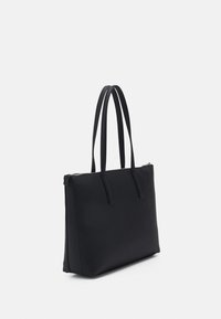 Musta kantokassi, jossa on sileä, mattapinta; varustettu kahdella pitkällä kahvalla ja vetoketjullisella ylhäällä. Suorakaiteen muotoinen ja minimalistinen muotoilu.