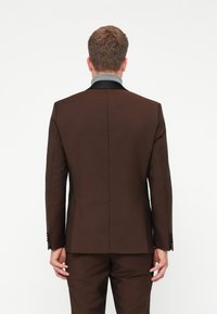 Bărbat îmbrăcat într-un costum maro închis și cămașă gri, stând cu spatele la cameră, pe un fundal alb simplu.