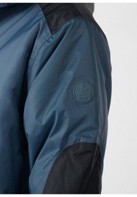 Una chaqueta azul ligera que presenta un hombro negro oscuro, tela texturizada y un parche de logo en relieve en la parte superior del brazo.