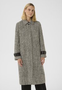 Femme portant un long manteau en chevron noir et gris avec des détails en cuir noir aux poignets, se tenant devant un fond clair.