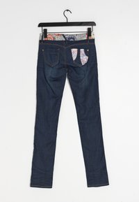 Jean bleu foncé avec deux poches arrière, une poche partiellement recouverte de tissu à motifs, suspendu à un cintre noir sur un fond blanc.