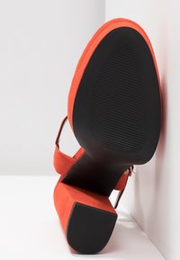 Punaju suede blok-topukna obuća postavljena pod kutom kako bi se prikazala crna gumena potplata s teksturiranim držačem i kopčom oko gležnja na bijeloj pozadini.