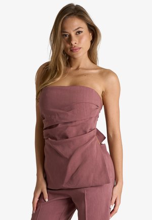 Donna con lunghi capelli biondi che indossa un top beige-mauve senza spalline e pantaloni coordinati, in piedi davanti a uno sfondo bianco semplice.