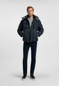 Mörkblå puffjacka med dragkedja framtill, stora fickor och huva, bärs över en grå tröja och matchas med mörka byxor och svarta sneakers.