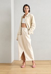 Chaqueta de denim beige, top corto blanco, falda beige hasta los tobillos con una abertura en el frente y zapatos de tacón rosa. Gafas con un tinte azul claro.