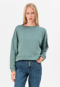 Sweatshirt verde scurt cu un croi relaxat, guler larg și umeri lăsați, asortat cu blugi albaștri cu talie înaltă. Textură netedă.