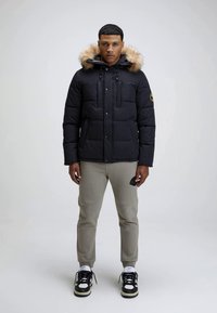 Junger Mann steht in einer schwarzen Daunenjacke mit pelzbesetzter Kapuze, hellgrauen Jogginghosen, weißen Socken und schwarz-weißen Turnschuhen vor einem einfarbigen Hintergrund.