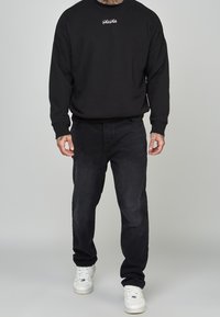 Felpa nera con logo bianco, abbinata a jeans scuri e sneakers bianche. Outfit casual caratterizzato da una vestibilità comoda e design semplice.