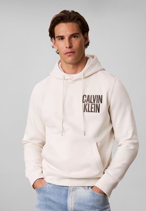 Jeune homme portant un sweat à capuche blanc Calvin Klein, les mains dans les poches, et un jean bleu clair, debout devant un fond gris uni.