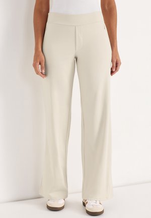 Beige wijde broek met voorzakken met rits, gedragen met witte en beige sneakers, getoond van taille tot voeten tegen een witte achtergrond.