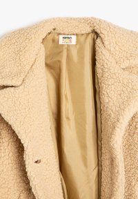 Abrigo de forro polar beige con un exterior texturizado, que presenta un interior suave en dorado. Incluye una etiqueta con información de tamaño y marca en su interior.