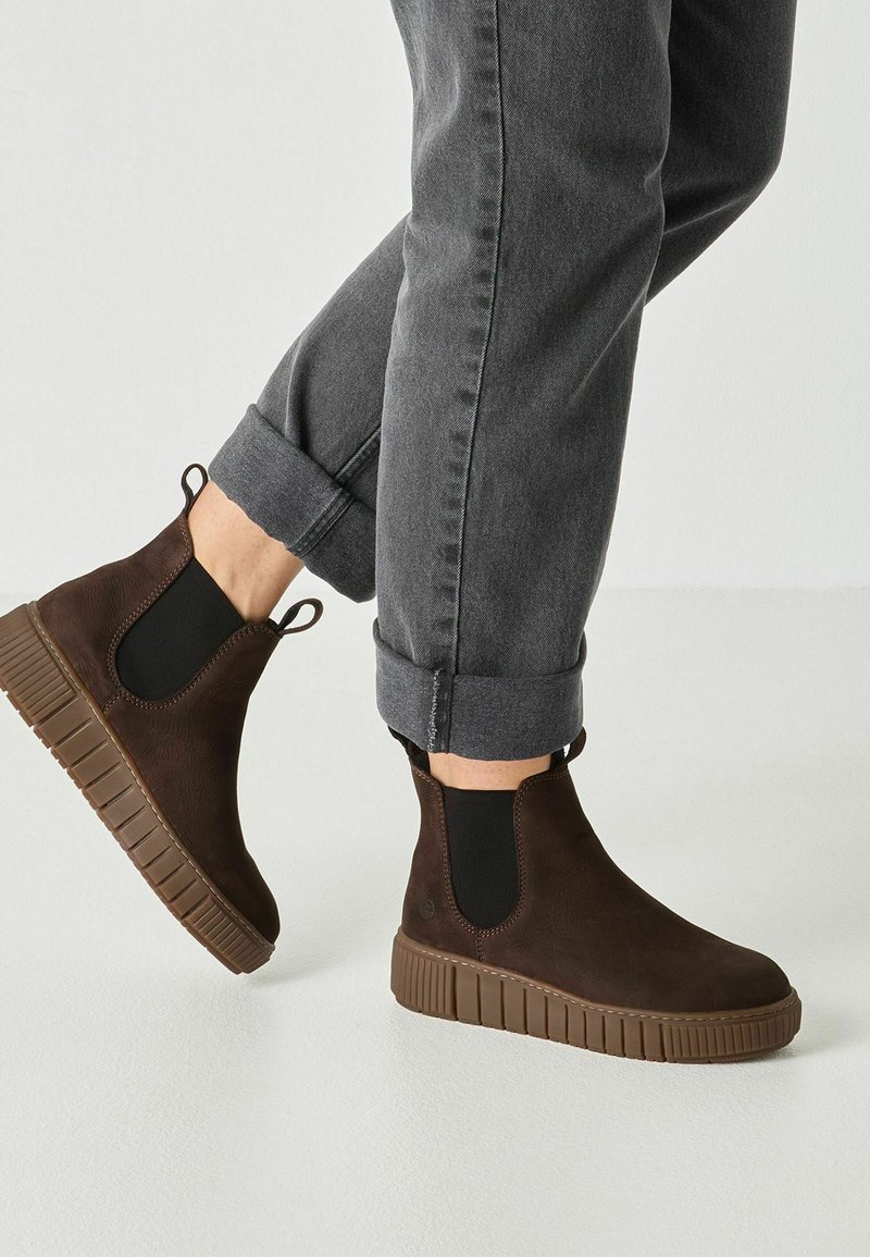 Personne portant des bottines basses marron foncé à enfiler avec des semelles épaisses en caoutchouc et un jean gris foncé retroussé sur un fond blanc uni.