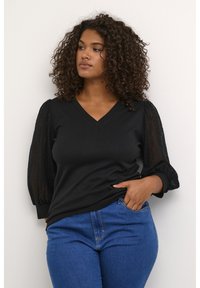 Schwarzes V-Ausschnitt-Top mit strukturierten Spitzen-Puffärmeln. Der Körper besteht aus einem einfarbigen schwarzen Stoff, kombiniert mit blauen Jeans.