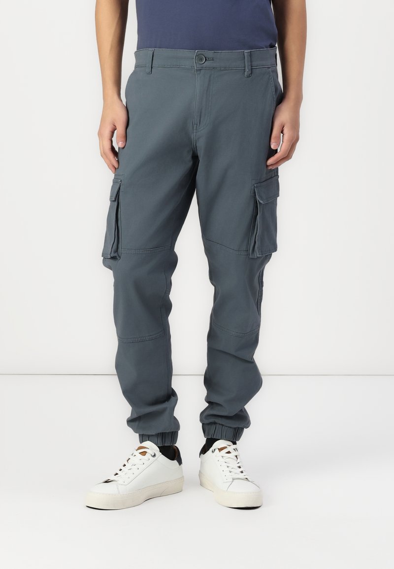 Spodnie cargo z ciemnoszarej tkaniny, z bocznymi kieszeniami, elastycznymi mankietami i prostymi nogawkami, noszone z białymi trampkami.