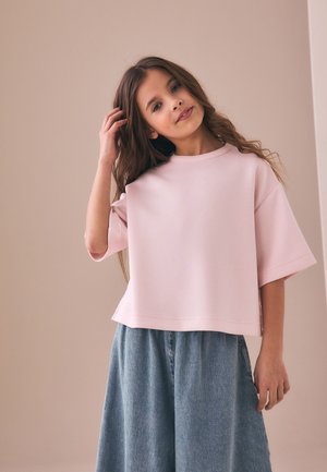 Junges Mädchen mit langen braunen Haaren, das ein locker sitzendes blassrosa Oberteil und blaue weite Hosen trägt, steht vor einem beigen Hintergrund.
