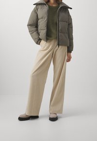 Person in olivgrünem Daunenjacke, grünem Strickpullover, beigen weitgeschnittenen Cordhose und beigen Stiefeln mit schwarzen Sohlen vor einem weißen Hintergrund.