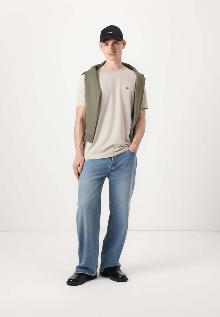 T-shirt beige clair avec un petit logo, pull vert drapé sur les épaules, jeans bleu clair, et casquette et chaussures noires. Coupe décontractée classique.