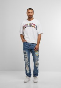 Bărbat stând cu o mână în buzunar, purtând un tricou alb cu logo-ul Colucci, blugi albaștri rupți cu petice și adidași albi, pe un fundal simplu.