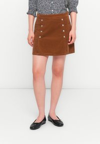 Brown corduroy mini skirt with silver button accents and side pockets, straight cut and textured fabric, paired with black ballet flats.