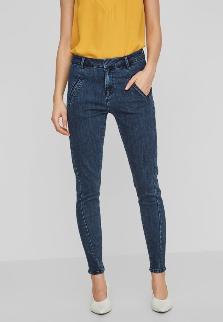 Jeans ajustados de denim azul oscuro, con bolsillos frontales y cintura de altura media. Combinados con una blusa amarilla y tacones blancos.