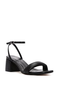 Sandale en cuir noir avec un talon épais et carré, bout ouvert, et une fine bride de cheville maintenue par une boucle métallique. Texture lisse.
