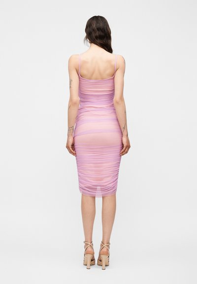 Γυναίκα με μακριά σκούρα μαλλιά φοράει ένα διάφανο λιλά midi φόρεμα με ραφές και λεπτές τιράντες, καθώς και μπεζ ψηλοτάκουνα σανδάλια, στέκεται μπροστά σε λευκό φόντο.