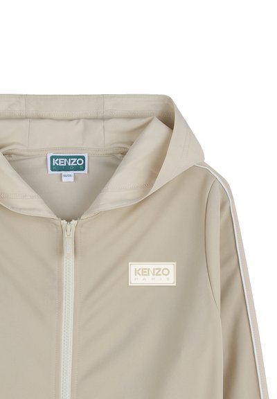 Beige gewatteerd jack van gladde stof met een centrale ritssluiting, zijstrepen en een wit logo-embleem op de borst.