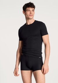 T-shirt nera aderente con maniche corte abbinata a boxer neri coordinati. Entrambi realizzati in un tessuto liscio ed elasticizzato.