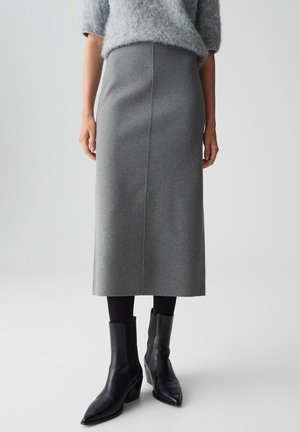 Kvinde iført en grå midikjol med højtaljet talje, sorte strømpebukser, sorte spidse ankelstøvler og en lysegrå, blød sweater.