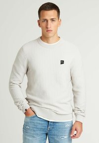 Hellgrauer, strukturierter Sweatshirt mit einem gerippten Rundhalsausschnitt und Bündchen. Verfügt über ein kleines schwarzes Logo-Patch auf der linken Brust. Trägt man über hellblauen Jeans.