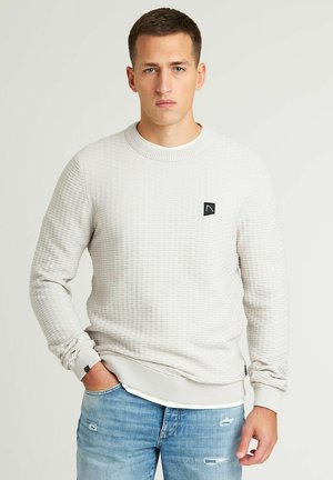Sweatshirt texturé gris clair avec un col rond et des bords côtelés. Présente un petit patch logo noir sur la poitrine gauche. Porté sur un jean bleu clair.