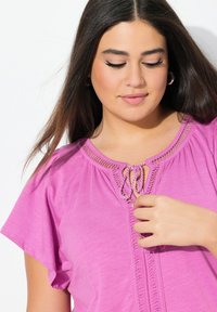 Růžový top s krátkým rukávem a kulatým výstřihem, který má zaplétanou šňůrku a strukturované detaily okolo výstřihu a vpředu.