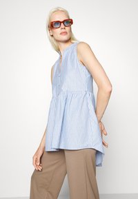 Blusa sem mangas azul e branca com riscas, com gola de botões e parte inferior larga. Combinada com calças largas castanhas, acentuadas por óculos de sol laranja.